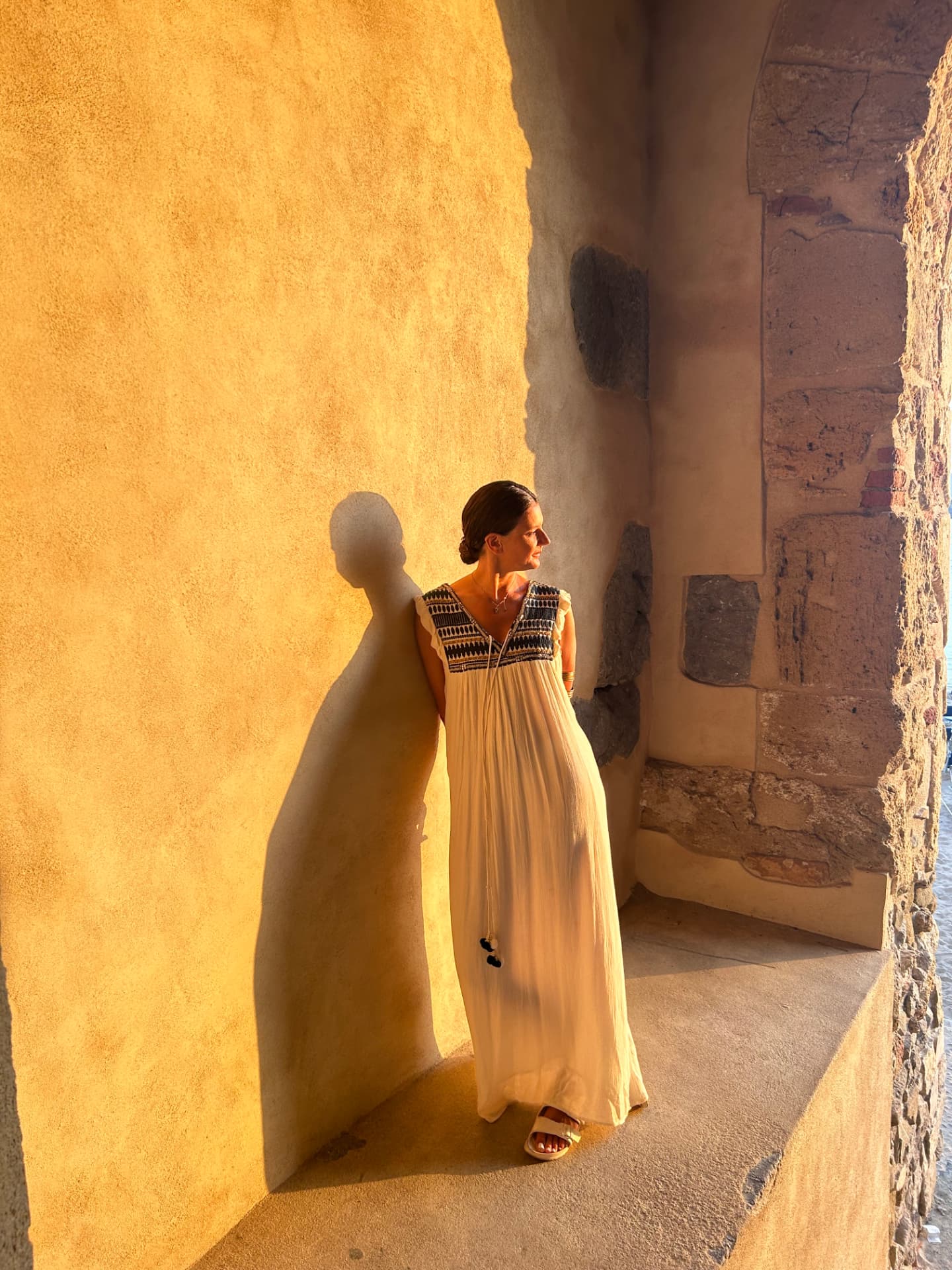 Zoë Hearne in a white dress, leaning against a sunlit ochre wall